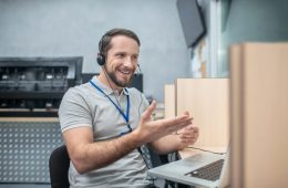 Work, emotion. Young adult smiling gesturing man in headphones near laptop at work place in room